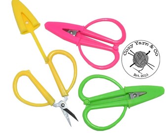 May include: Three pairs of small craft scissors with plastic handles in yellow, pink, and green, each featuring a matching protective cap. A circular logo with the text Cozy Yarn & Co Est. 2013 and a yarn ball illustration is included.