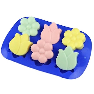 May include: A blue silicone mold with six cavities shaped like flowers and tulips. The mold is filled with colorful soap bars in shades of pink, yellow, and green.