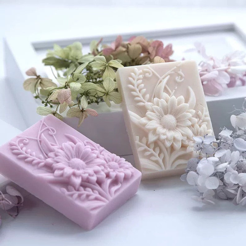 May include: Two rectangular bars of soap with floral designs. The soap on the left is a light purple color and the soap on the right is a light beige color.