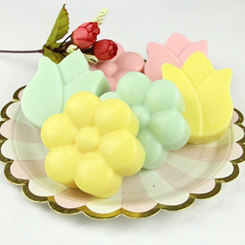 May include: Five colorful handmade soap bars shaped like flowers and leaves. The soaps are arranged on a pink and white striped paper plate with a gold scalloped edge.