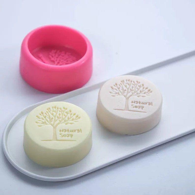 May include: Two round, white soap bars with a tree design and the words "Natural Soap" imprinted on them. A pink silicone mould with the same tree design is also shown.