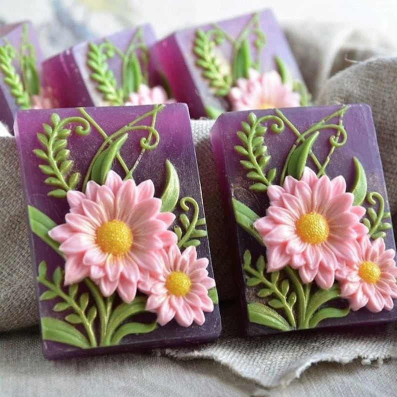 May include: Purple and green floral soap bars with pink flowers and green leaves. The soap bars are arranged in a row on a light brown fabric surface.