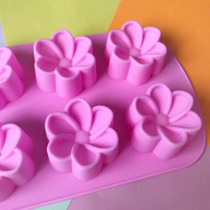 May include: Pink silicone mould with six flower-shaped cavities. The mould is designed for making small cakes, chocolates, or other treats.