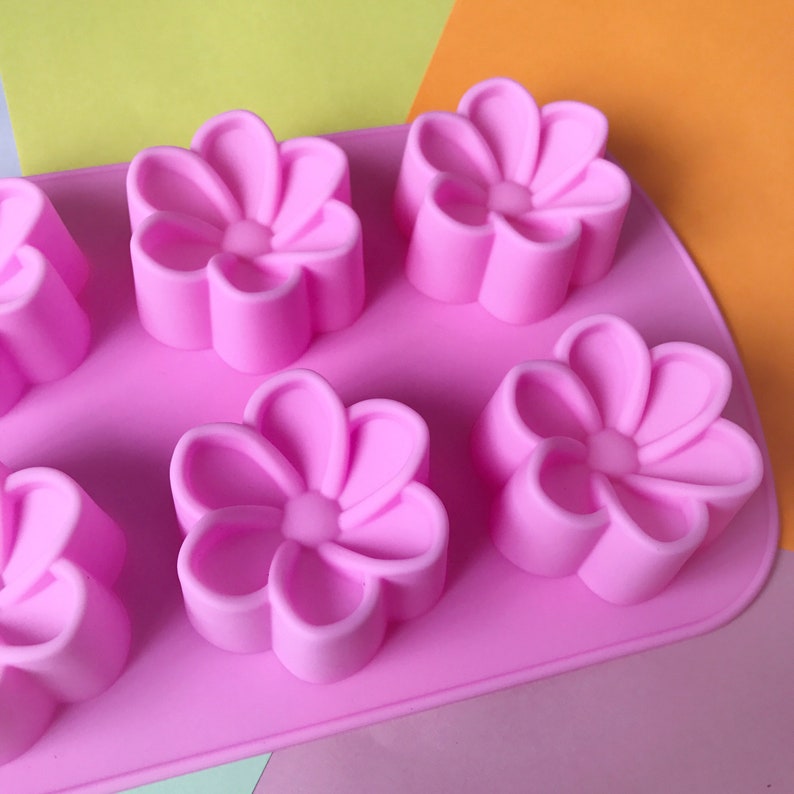 May include: Pink silicone mould with six flower-shaped cavities. The mould is designed for making small cakes, chocolates, or other treats.