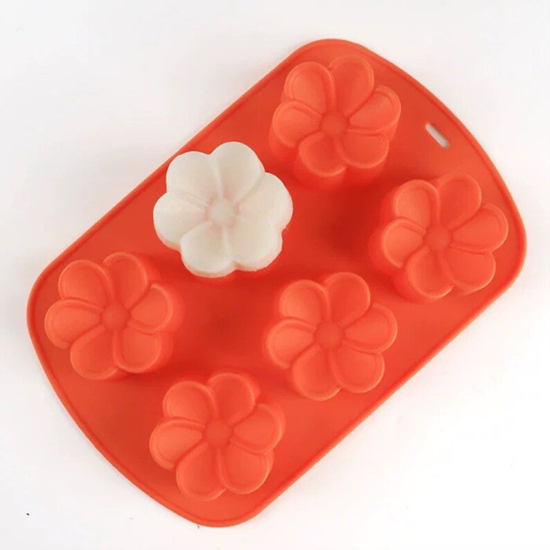 May include: Orange silicone mould with six flower-shaped cavities. One cavity is filled with a white soap bar.