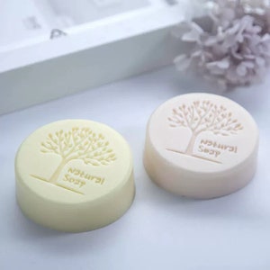 May include: Two round bars of natural soap with a tree design and the words "Natural Soap" printed on them. One bar is pale yellow and the other is pale beige.