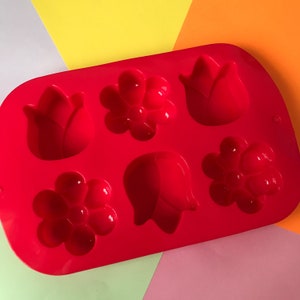 May include: Red silicone mold with six cavities shaped like flowers and tulips. The mold is designed for making small cakes, candies, or other treats.