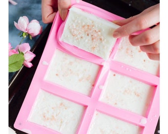 May include: Pink silicone mould with six rectangular cavities filled with white soap with pink salt. A hand is pressing a piece of soap into one of the cavities.