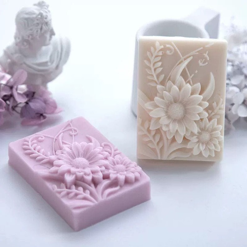 May include: Two rectangular bars of soap with floral designs. One bar is white with a raised design of white flowers and leaves. The other bar is purple with a raised design of purple flowers and leaves.