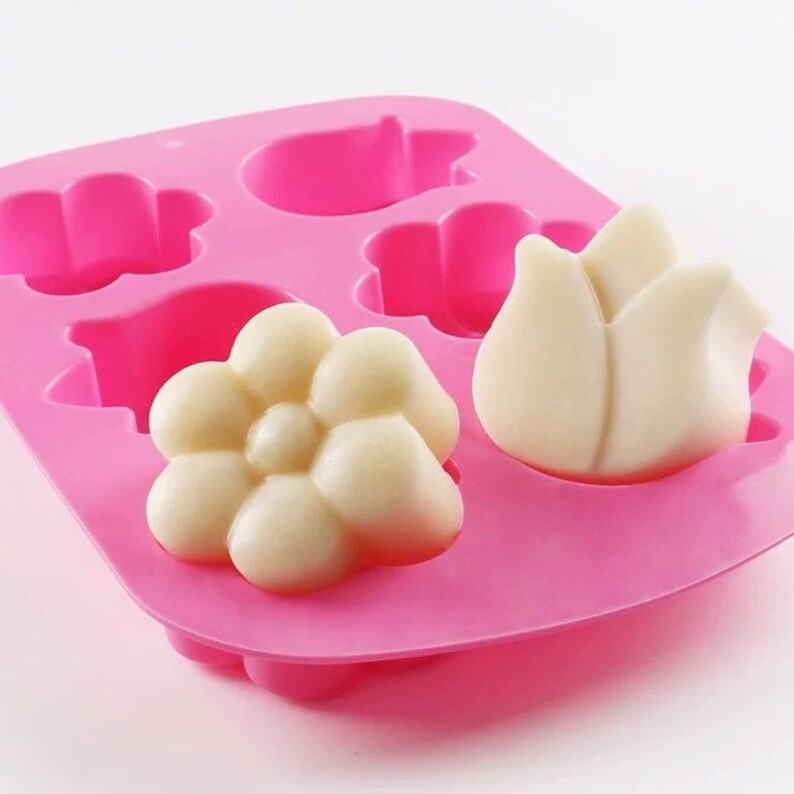 May include: Pink silicone mold with six cavities shaped like flowers and a tulip. The mold is filled with white soap in the shape of a flower and a tulip.