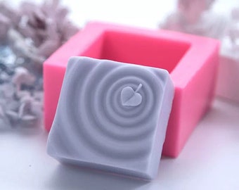 May include: A light purple soap bar with a swirl pattern and a small leaf design in the centre. The soap bar is in a pink silicone mould.
