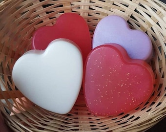 May include: A woven basket holds five heart-shaped soaps in shades of red, pink, and white. One soap is white, two are red, one is pink with glitter, and one is lavender. The basket is made of woven, light brown material.