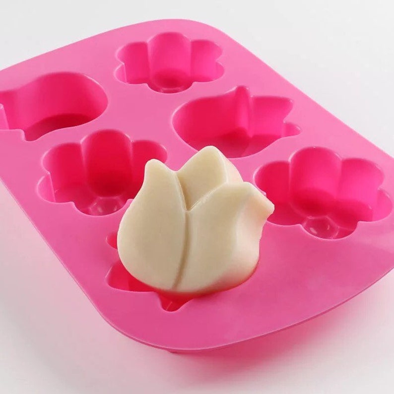 May include: Pink silicone mold with six cavities shaped like flowers and tulips. A white soap bar shaped like a tulip is in one of the cavities.