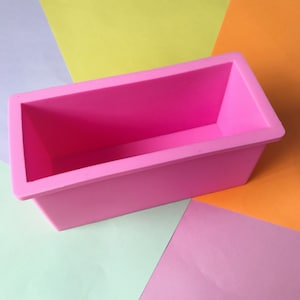 Puede incluir: Un molde rectangular de silicona rosa brillante para hacer jabón o hornear. El molde tiene un diseño sencillo de tapa abierta con lados ligeramente inclinados. El fondo es una colorida disposición de papel amarillo, naranja, rosa y verde.