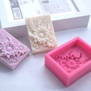 May include: Three silicone molds for making soap bars. The molds are rectangular and feature a raised design of flowers and leaves. One mold is white, one is purple, and one is pink.