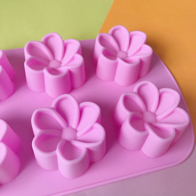 May include: Pink silicone mould with six flower-shaped cavities. The mould is designed for making small cakes, chocolates, or other treats.