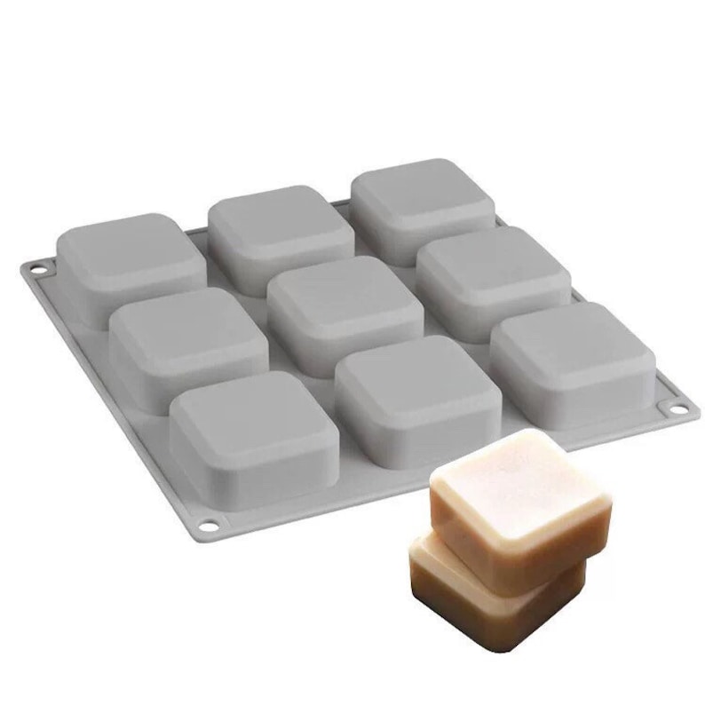 May include: Gray silicone mold with 12 square cavities for making soap bars. Two bars of soap are shown next to the mold.