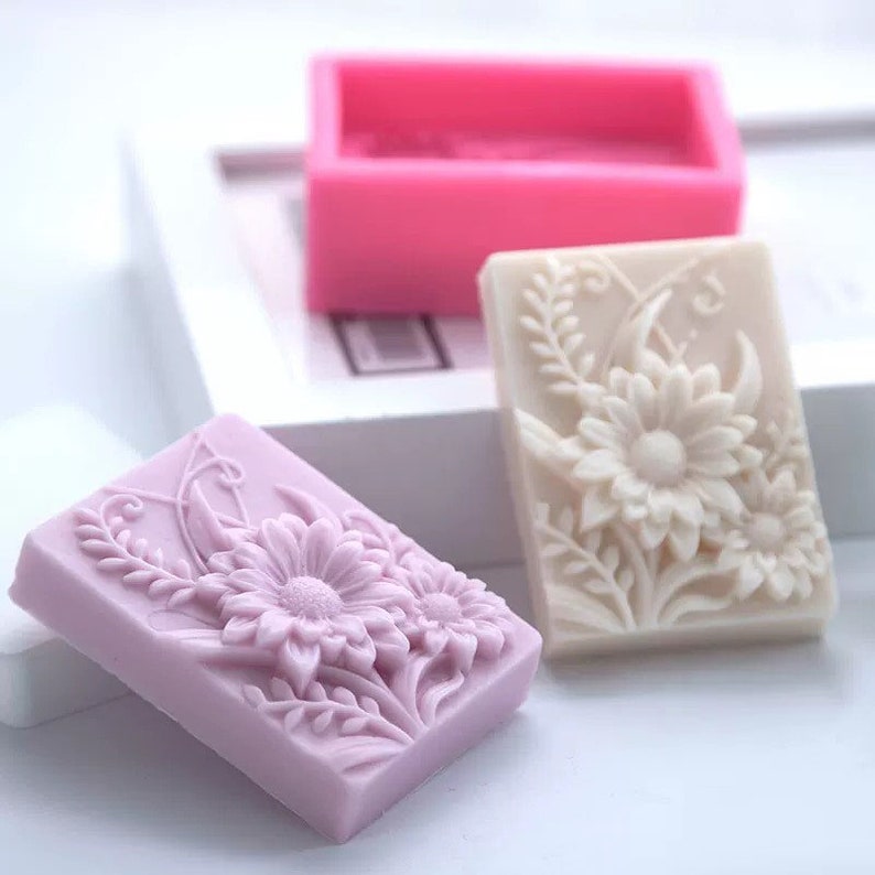 May include: Three rectangular silicone molds for making soap. The molds are in pink, white, and purple. Each mold features a detailed floral design with a raised pattern of flowers and leaves.