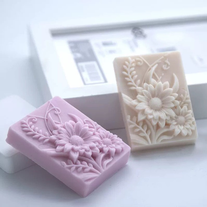 May include: Two rectangular soap bars with floral designs. One bar is light pink with a detailed design of three flowers and leaves. The other bar is off-white with a similar design of three flowers and leaves.