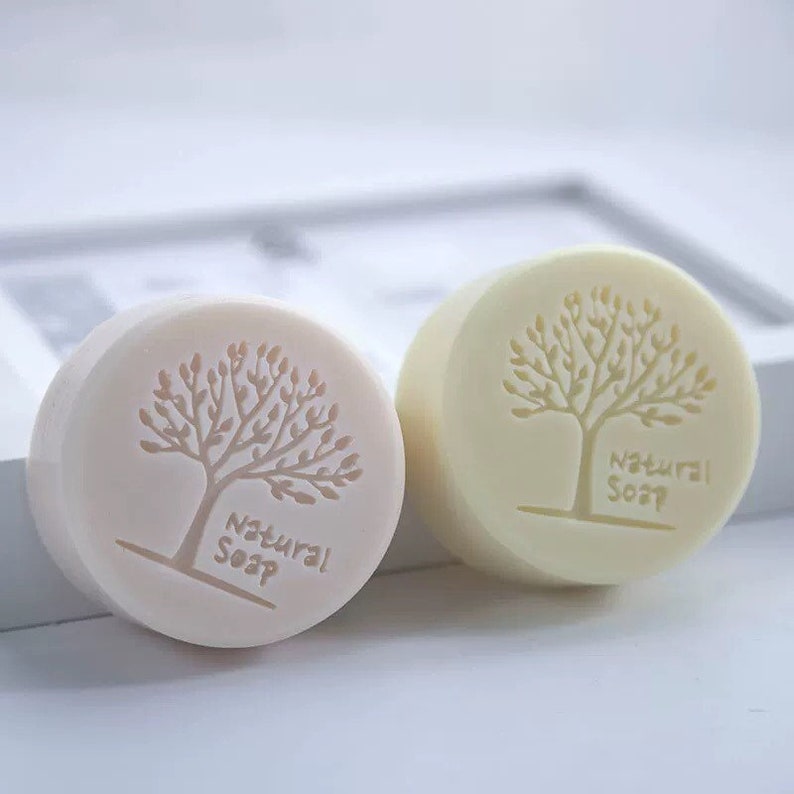 May include: Two round bars of soap with a tree design and the words "Natural Soap" on them. One bar is a light beige colour and the other is a light yellow colour.