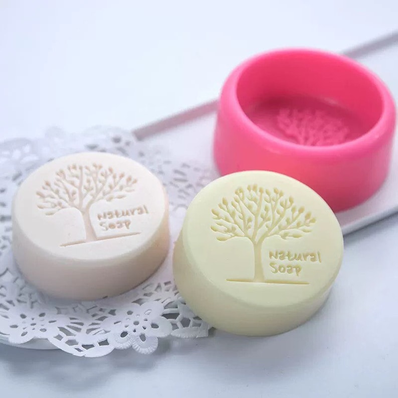 May include: Two round, white soap bars with a tree and "Natural Soap" text embossed on the top. A pink silicone mould for making soap is also shown.