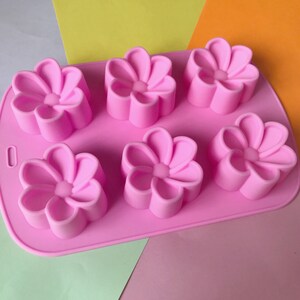 May include: Pink silicone mould with six flower-shaped cavities. The mould is designed for making small cakes, soaps, or other crafts.