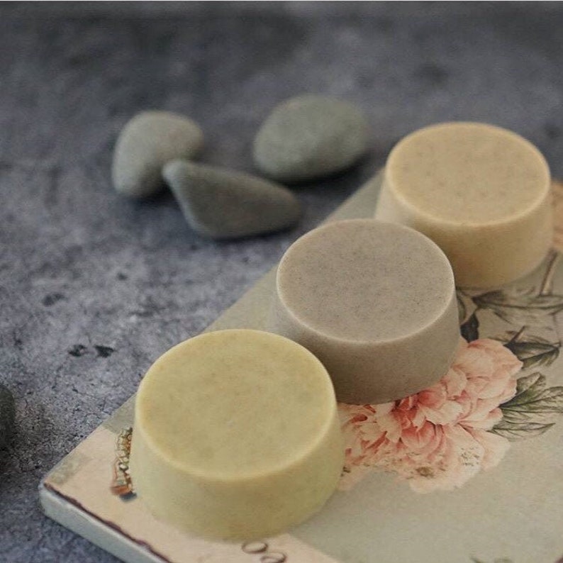 May include: Three round bars of soap, one is light green, one is light brown, and one is a darker brown. The soap bars are sitting on a floral patterned surface.