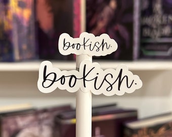 Può includere: Due adesivi bianchi con la parola "bookish" scritta in corsivo nero. Gli adesivi sono attaccati a una penna bianca, che si trova davanti a una libreria.