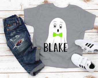 May include: A gray t-shirt with a white ghost wearing a green bow tie. The ghost has two large eyes and a surprised expression. The text "BLAKE" is printed below the ghost.