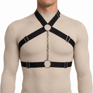 May include: A black elastic chest harness featuring silver-toned metal hardware, including adjustable sliders, two circular rings, and a vertical metal chain connecting the neck and waist straps, worn over a beige long-sleeved shirt.