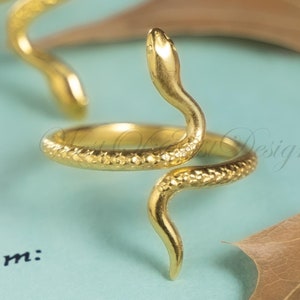 May include: Gold-colored snake ring with textured scales, coiled around the finger. The ring is set against a light blue background with the words "To:" and "From:" visible. A dried leaf adds a natural element.