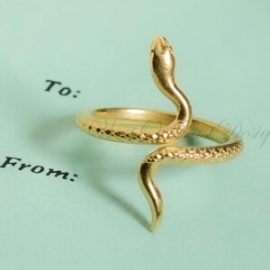 May include: A gold-colored snake ring with a textured surface. The snake's head is raised, and the body coils around the finger. The ring is set against a light blue background with the words "To:" and "From:" printed in black.