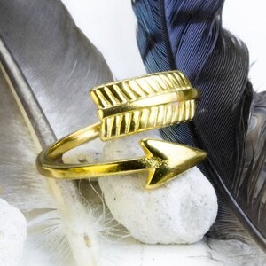 May include: Two gold arrow rings with a simple design. The rings are on a white surface with feathers in the background.
