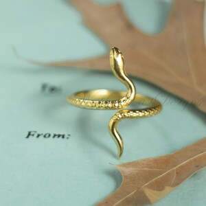 May include: A gold-colored snake ring with a textured surface. The snake's head is raised, and its body wraps around the finger. The ring is set against a light blue background with a leaf.