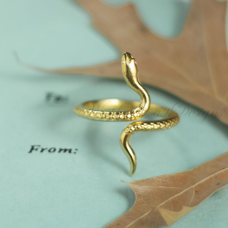 May include: A gold-colored snake ring with a textured surface. The snake's head is raised, and its body wraps around the finger. The ring is set against a light blue background with a leaf.