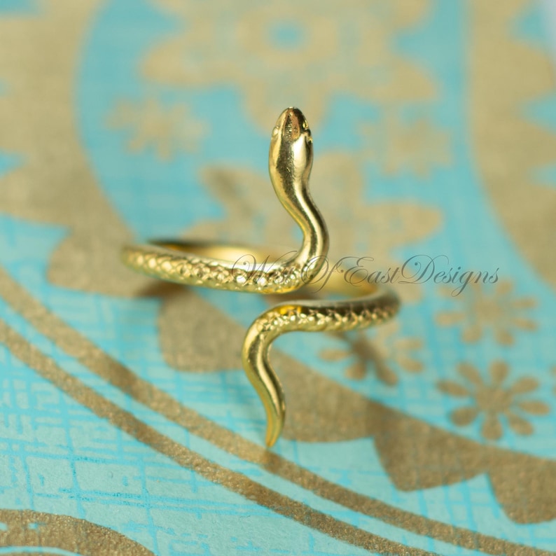 May include: A gold-colored snake ring with a detailed design. The snake's head is raised, and the body wraps around the finger. The ring is set against a blue and gold patterned background.