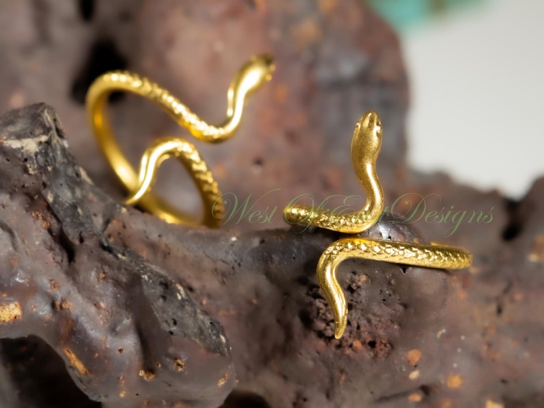 May include: Two gold-colored snake rings. The rings are coiled, with the snake heads raised. The rings are on a dark, textured surface. The rings have a shiny, metallic finish. The text "West Coast Designs" is visible in the background.