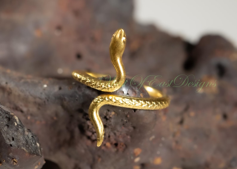 May include: A gold-colored snake ring with a textured, scale-like pattern. The snake's head is raised, and the body wraps around the finger. The ring is set against a dark, textured background.