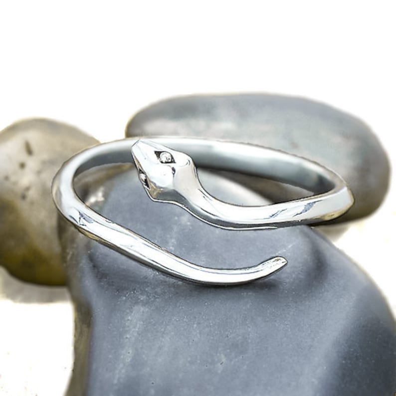 May include: A silver-colored ring in the shape of a snake, coiled around a gray stone. The snake's head is detailed with small, dark accents for eyes. The ring's design is smooth and polished, reflecting light.