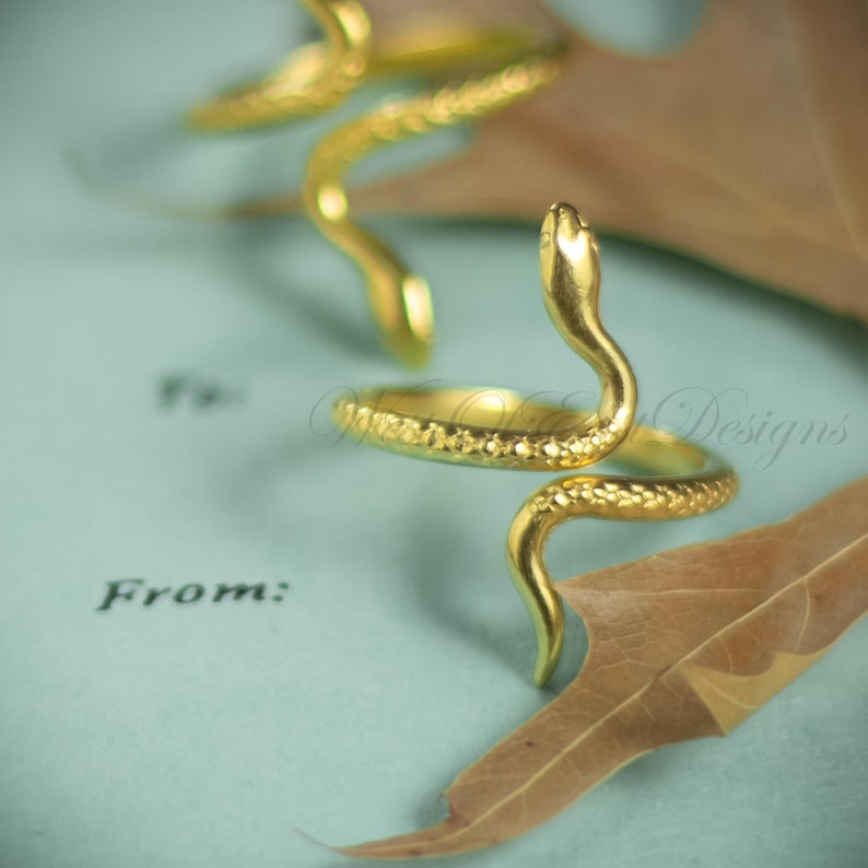 May include: A gold-colored snake ring with textured detailing. The snake's head is raised, and the body coils around the finger. The ring is set against a light blue background with a dried leaf.