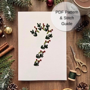Puede incluir: Una tarjeta rectangular blanca con una forma de bastón de caramelo bordada con hojas verde oscuro y pequeñas bayas rojas. El texto PDF Pattern & Stitch Guide está impreso en negro sobre la tarjeta, rodeada de decoraciones navideñas.