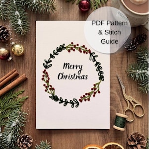 Może przedstawiać: Biała prostokątna kartka z wyhaftowanym wieńcem z zielonych liści i czerwonych jagód otaczającym czarny napis Merry Christmas. Kartka jest oznaczona jako PDF Pattern & Stitch Guide i otoczona świątecznymi dekoracjami.