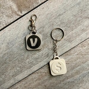 May include: Two keychains with square pendants. One pendant is silver with a black circle and a white "V" inside. The other is white with a raised dollar sign. Both have metal key rings and clasps.