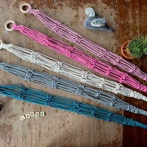 May include: Five macrame plant hangers in different colours, including teal, grey, white, pink, and purple. The hangers are made with cotton cord and have wooden rings at the top. The word "LARGE" is spelled out in wooden letter tiles.