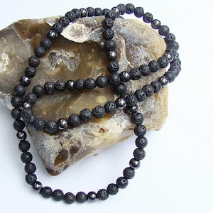 Może przedstawiać: Naszyjnik z czarnych koralików z lawy wulkanicznej i hematytu. Naszyjnik wykonany jest z małych, okrągłych koralików i leży na kawałku brązowego kamienia.