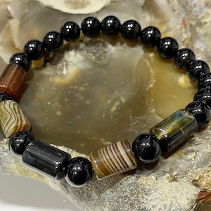 May include: Black onyx bead bracelet with four brown and white agate beads. The bracelet is on a brown and gray stone background.