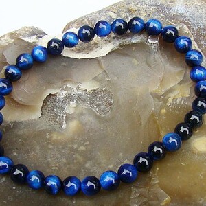 Puede incluir: Una pulsera de cuentas con cuentas redondas pulidas en tonos azul oscuro y negro. Las cuentas tienen un acabado brillante y están ensartadas para formar una banda flexible. La pulsera se muestra sobre una superficie clara y texturizada.