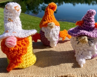 May include: Three crocheted gnomes, one with a white hat and orange and yellow beard, one with an orange hat and purple and white beard, and one with a purple hat and white beard.