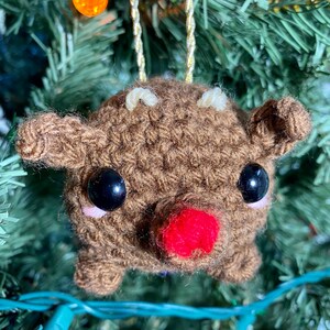 May include: A handmade brown crochet reindeer Christmas ornament. The reindeer has black eyes, a red nose, and small white antlers. It is suspended by a gold cord and is displayed on a Christmas tree with colorful lights.