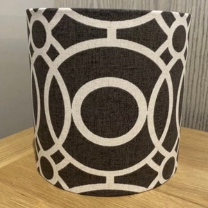 May include: Cylindrical lampshade featuring a dark grey backdrop and a white geometric design. The pattern is made up of interconnected circles and lines, creating a modern aesthetic. The lampshade is placed on a light-coloured wooden surface.